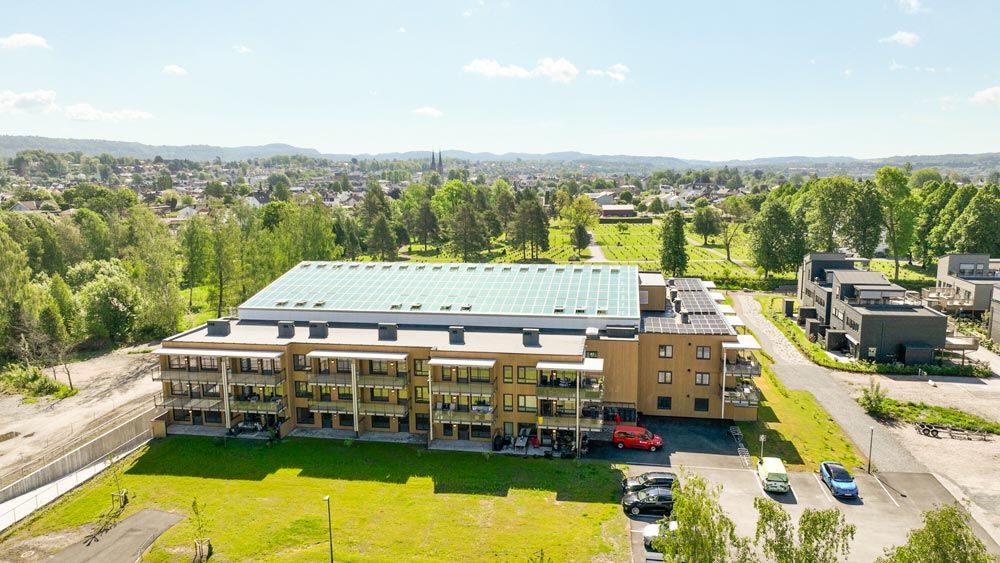 Fugleperspektiv av Bovieran Falkum leilighetsbygg i Skien med innglasset vinterhage, solceller på taket, grøntområder og parkeringsplass i et naturskjønt boligområde.